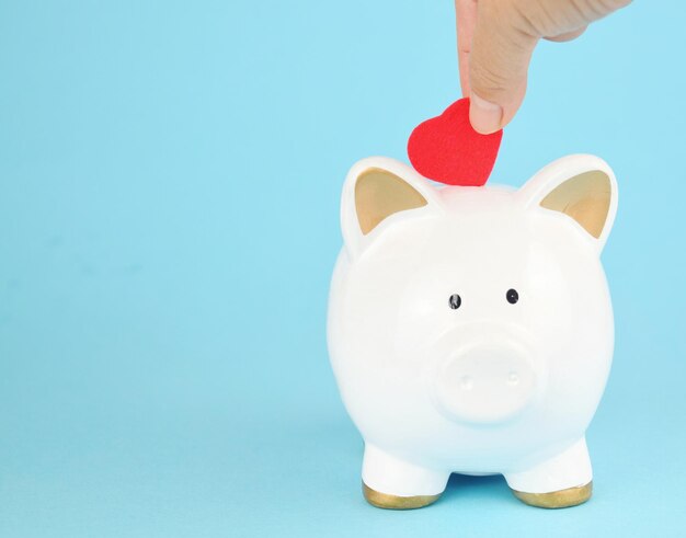 Person hand putting heart shape paper in piggy bank against blue background