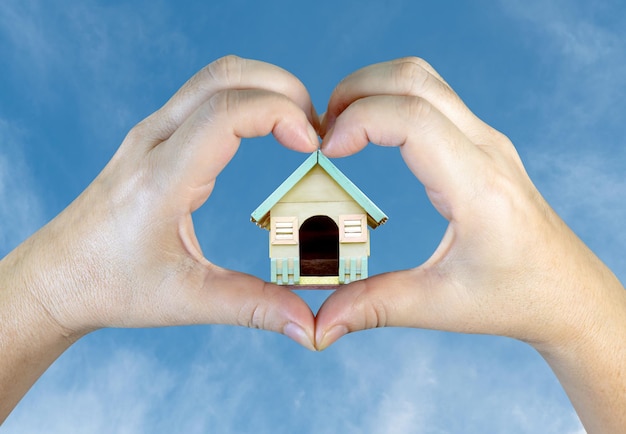 person hands making a heart shape with wooden house on sky background
