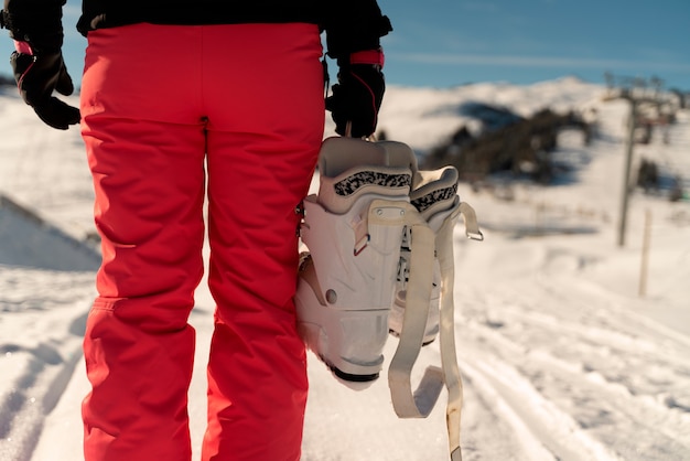 Person on her back wearing a pink ski pant holding a pair of ski boots in a ski resort in the Alps