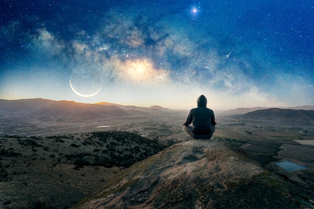 은하수와 달을 배경으로 밤에 야외에서 명상하거나 기도하는 언덕 위에 있는 사람