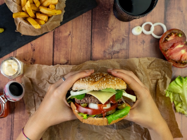 Person holding a burger with fries, onion rings and tomato on wooden table