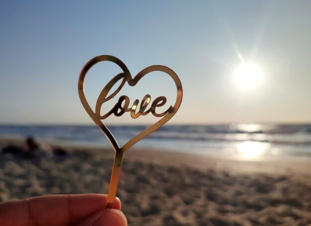 Person holding in fingers hand stick in shape golden heart and word love on background sea and sea