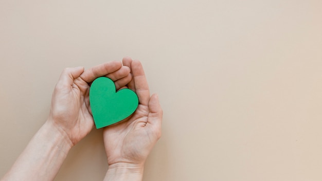 Person holding a green heart on beige background with copy space