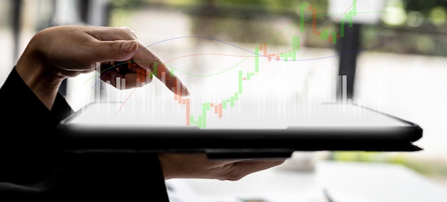 Человек, держащий планшет и указывающий на диаграмму, показывающую восходящий и нисходящий свечной график акций, графически отображаемый на экране планшета как восходящий тренд. Концепция инвестора акций.