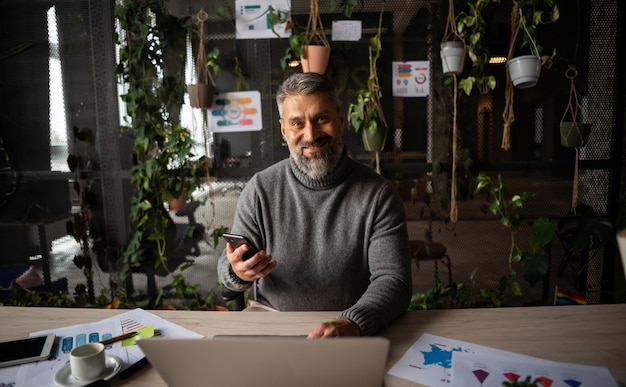 A person is seated at a wooden desk using a laptop while holding a cell phone nearby is a plant