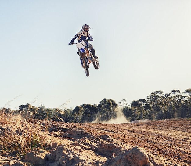 Photo person jump and motorbike of professional motorcyclist in the air for trick stunt or race on outdoor dirt track expert rider on bike or scrambler in dunes or extreme sport with blue sky background