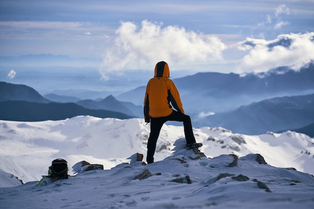 A person in an orange jacket stands on a snowy mountain top looking at the mountains.