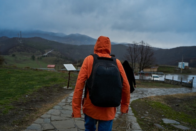 バックパックを背負ったオレンジ色のレインコートを着た人が山の雨の中を歩きます。