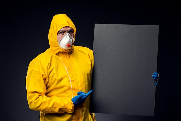 Человек в защитном костюме, очках и перчатках, изолированных на черном фоне, держит черную пустую доску с местом для текстового изображения Концепция пандемии