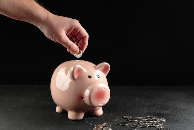 A person puts a coin in a piggy bank with a stack of coins next to it on a concrete table on a dark