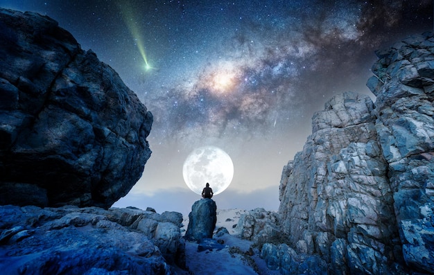 Person on the rock outdoors meditating or praying at night\
under the milky way and moon, back view