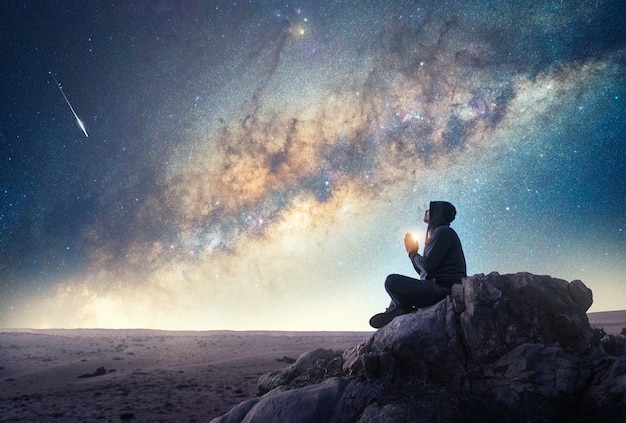 Person on the rock outdoors meditating or praying at night under the Milky Way and shooting star