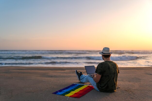 Person on the shore of the beach with a laptop and a rainbow flag looking at the sunset