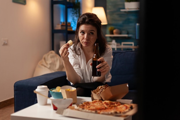 Person sitting on sofa looking worried at television while\
holding beer bottle and potato chip in home living room. woman\
office worker watching concerning tv news while having takeout\
dinner.