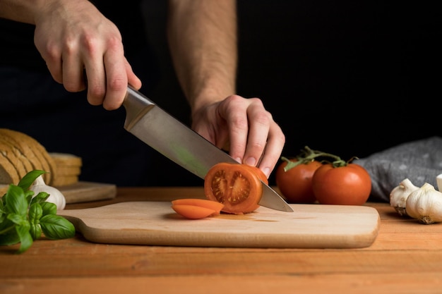 Человек, нарезающий помидоры на деревянной доске, готовит итальянскую брускетту на темном фоне
