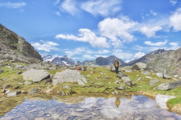 A person standing in front of a mountain lake with a blue sky and clouds in the background.