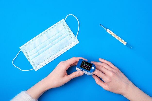 Person using pulse oximeter device on finger near to thermometer and medical mask, healthcare monitoring concept