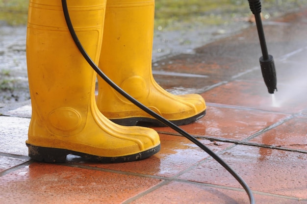 Person wearing yellow rubber boots with highpressure water nozzle cleaning the dirt in the tiles