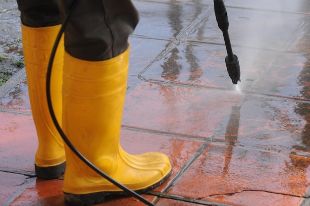 Person wearing yellow rubber boots with highpressure water nozzle cleaning the dirt in the tiles