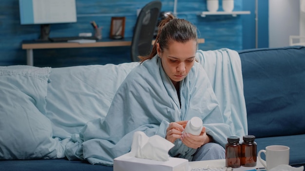 Person with cold reading label of bottle with capsules and pills against seasonal flu. Ill woman looking at jar with medicaments to cure virus disease for healthcare. Sick adult in blanket