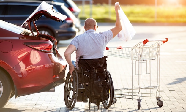 Человек с ограниченными физическими возможностями кладет покупки в багажник автомобиля на стоянке супермаркета.