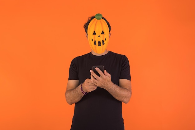 オレンジ色の背景に彼の携帯電話を見てハロウィーンを祝うカボチャのマスクを持つ人
