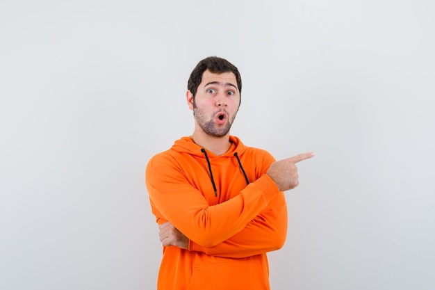 Photo personable man pointing left and surprised in orange hoodle and looking puzzled