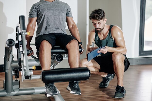 Personal trainer asking client to do exercise in leg extension machine and taking notes in document in clipboard