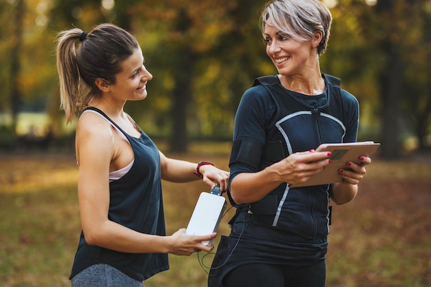 Personal trainer checking battery on a suit and teaching a smiling mature woman to working out with ems device in the city park.