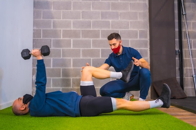 Personal trainer in de sportschool corrigeert de oefeningen van de atleet in de pandemie van het coronavirus een nieuw normaal Met beschermend gezichtsmasker