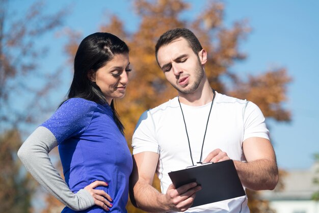 Personal Trainer Takes Notes While Young Woman Exercise in City Park Area  Training and Exercising for Endurance  Healthy Lifestyle Concept Outdoor