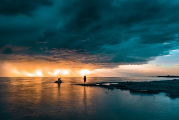 Persoon die aan de kust staat, omringd door de zee onder een stormachtige lucht tijdens een adembenemende zonsondergang