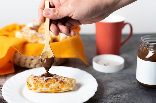 Persoon die chocolade op een bladerdeeggebak legt naast een mand vol snoep en een kopje koffie