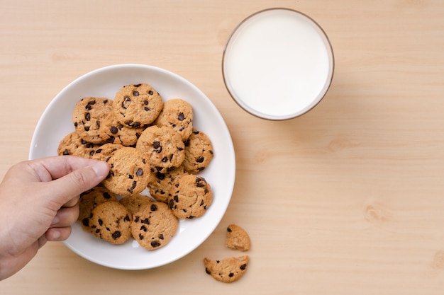 Foto persoon die een chocoladeschilferkoekje uit de schotel en melk op de houten lijst plukt