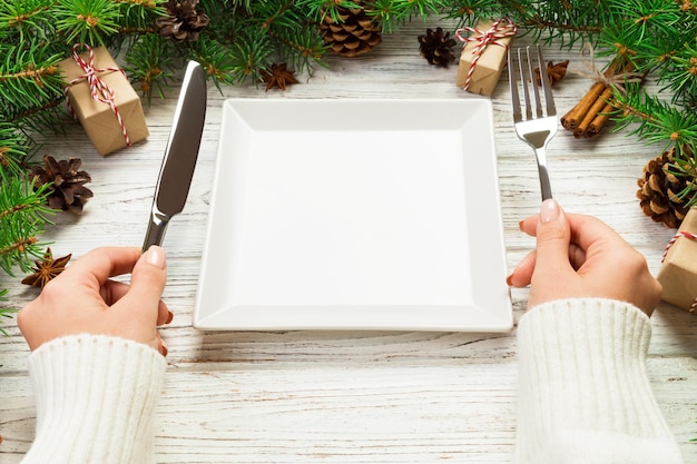 Perspectief weergave meisje houdt vork en mes in de hand en is klaar om te eten lege witte vierkante plaat op houten kerst achtergrond vakantie diner schotel concept met nieuwjaar decor
