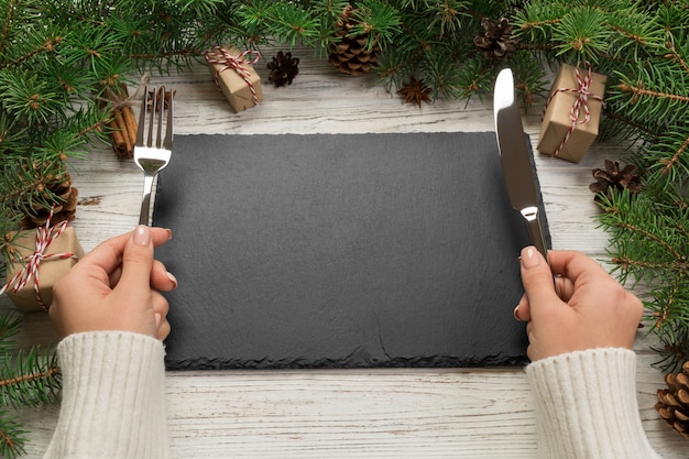 Perspective view girl holds fork and knife in hand and is ready to eat, Empty black slate rectangular plate on wooden christmas , holiday dinner dish concept with new year decor