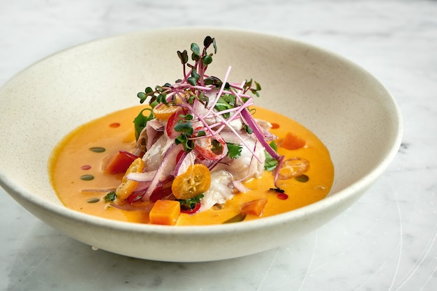 Peruvian cuisine dish - seabass ceviche with hot peppers, onions and yellow sauce, served in a white plate on a marble table. Restaurant seafood.