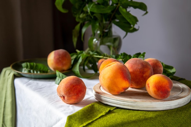 Perziken op tafel in de keuken bij het raam. Rustiek minimalisme eten perzik fruit met bladeren op een houten bord op tafelkleed. Oogst van rijpe sappige perziken. Stilleven Perziken fruit.