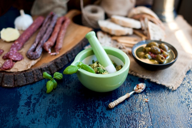 pestosaus in een groene kop en geassorteerde salami op een houten bord en olijven en blauw oppervlak