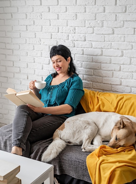 Foto cura degli animali domestici sorridente donna di mezza età che si diverte ad essere a casa e a leggere seduta sul divano