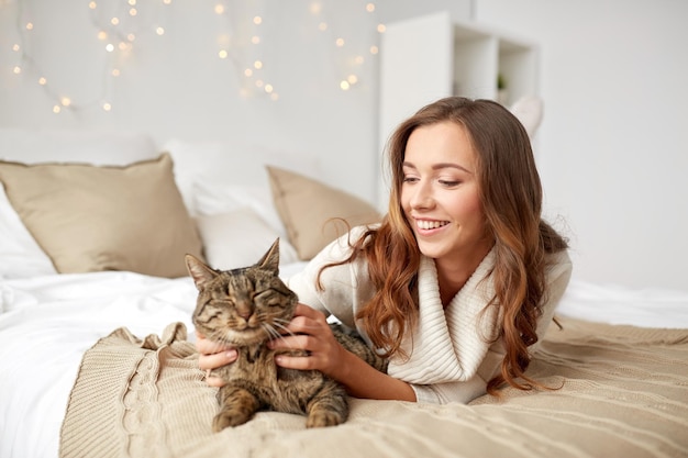 pets, comfort, christmas, winter and people concept - happy young woman with cat lying in bed at home