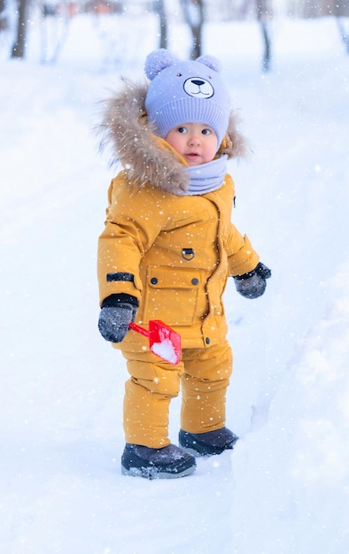 Peuter 15-20 maanden in gele warme kleren staat tussen sneeuwbanken in een winterpark met een speelgoedschop in zijn hand