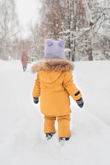 Peuter van 1217 maanden oud in gele warme kleding zorgt voor de vertrekkende moeder die in de sneeuw in een winterpark staat