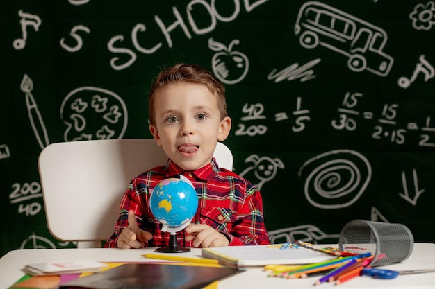 Peuterjongen die schoolthuiswerk maken. Schooljongen met blije gezichtsuitdrukking in de buurt van bureau met schoolbenodigdheden