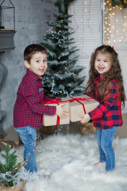 Peuterjongen en -meisje in rode shirts houden geschenkdozen bij de kerstboom in een licht kerstinterieur Leuke kerstkinderen