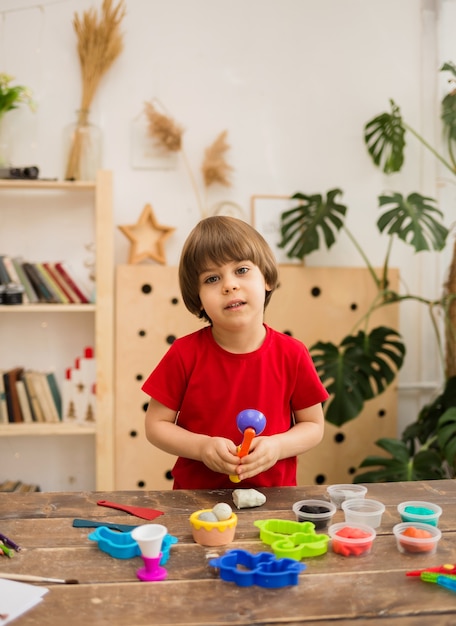 Peuterjongen in een rood T-shirt speelt met plasticine en vormen op houten tafel in de kamer. Ontwikkeling van fijne motoriek.