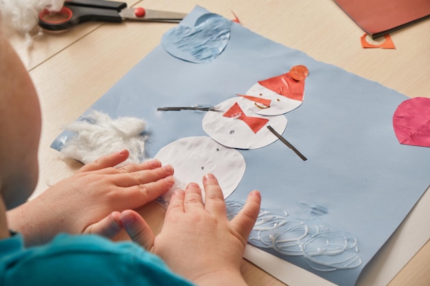 Peutermeisje in een blauw t-shirt maakt een applique terwijl ze aan tafel zit, papieren sneeuwpop