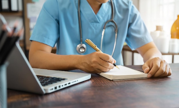 Pharmacist writing making notes in drug information using
laptop computer sitting at desk woman physician nurse or pharmacist
wearing white coat writing in paper notebook