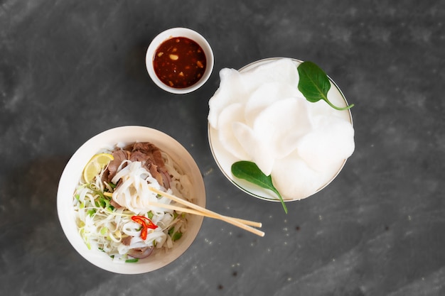 Pho bo Vietnamese noodle soup with beef and a traditional snack of rice chips and sesame sauce on a gray background