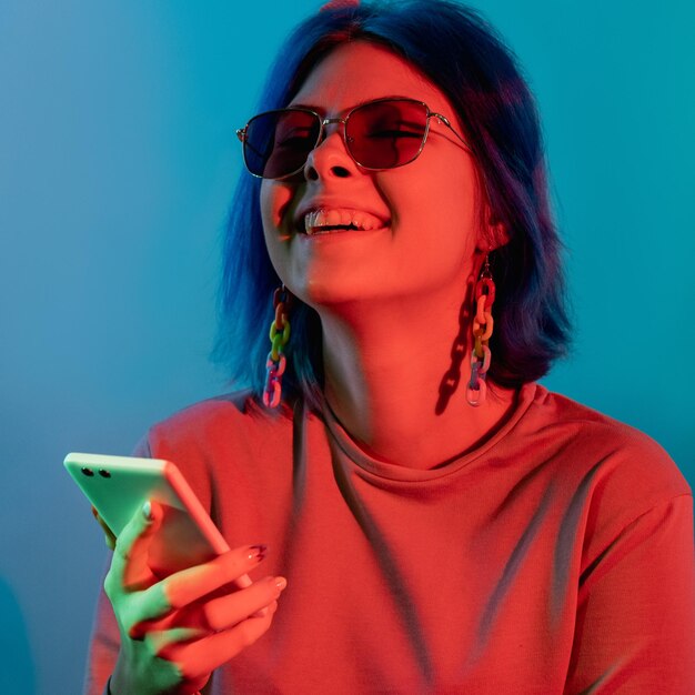 電話エンターテインメントミレニアルライフスタイルモバイル技術青い背景に分離された赤いネオン光でスマートフォンを使用してうれしそうな笑う女性